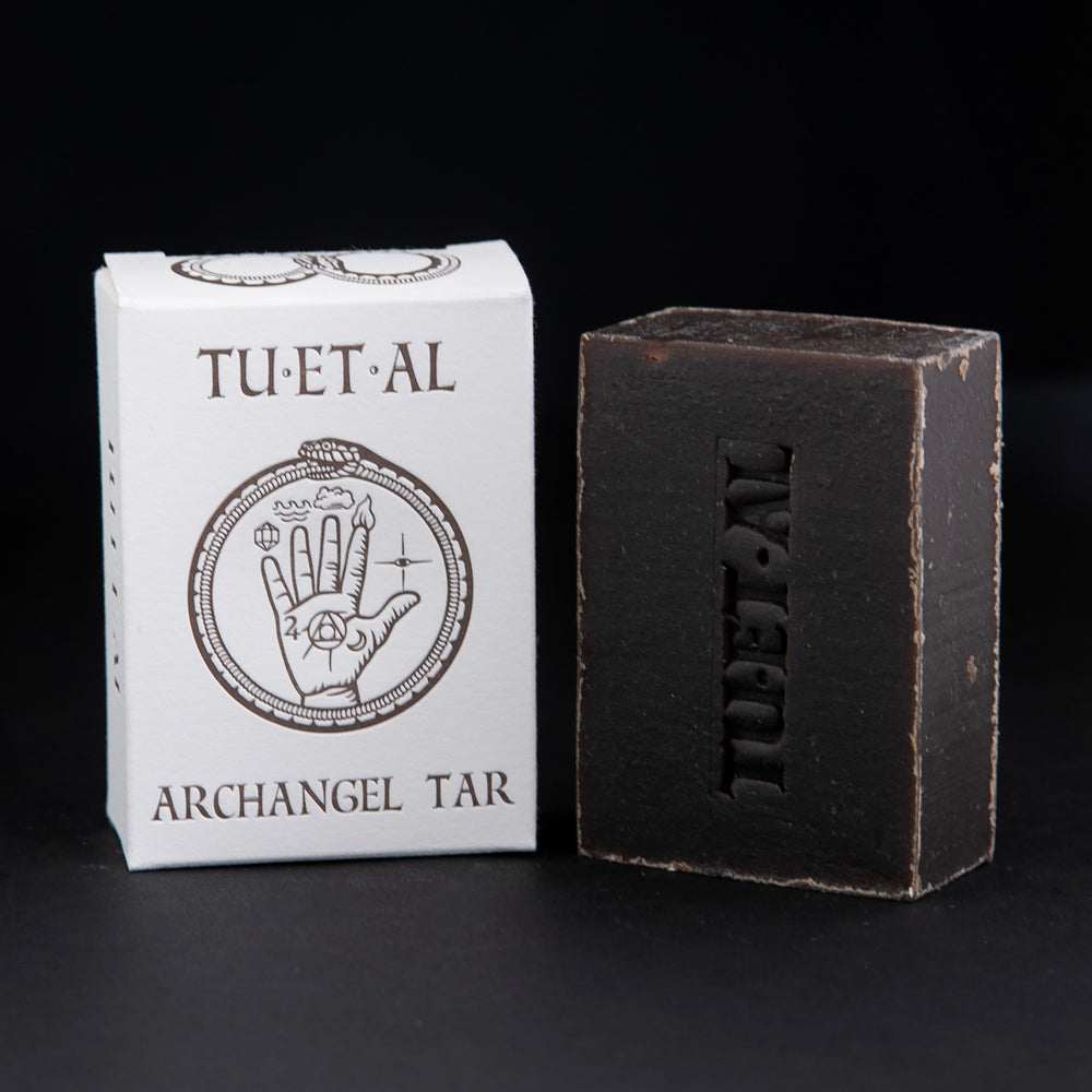 Box and bar of TU-ET-AL's "Archangel Tar" soap sitting against a black background. The bar of soap is brown.