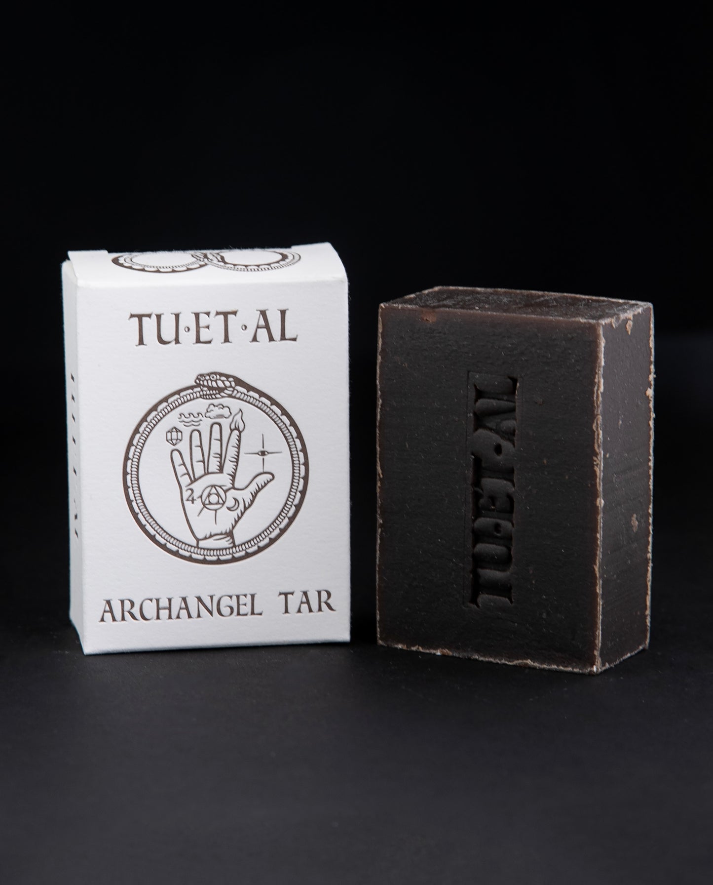 Box and bar of TU-ET-AL's "Archangel Tar" soap sitting against a black background. The bar of soap is brown.