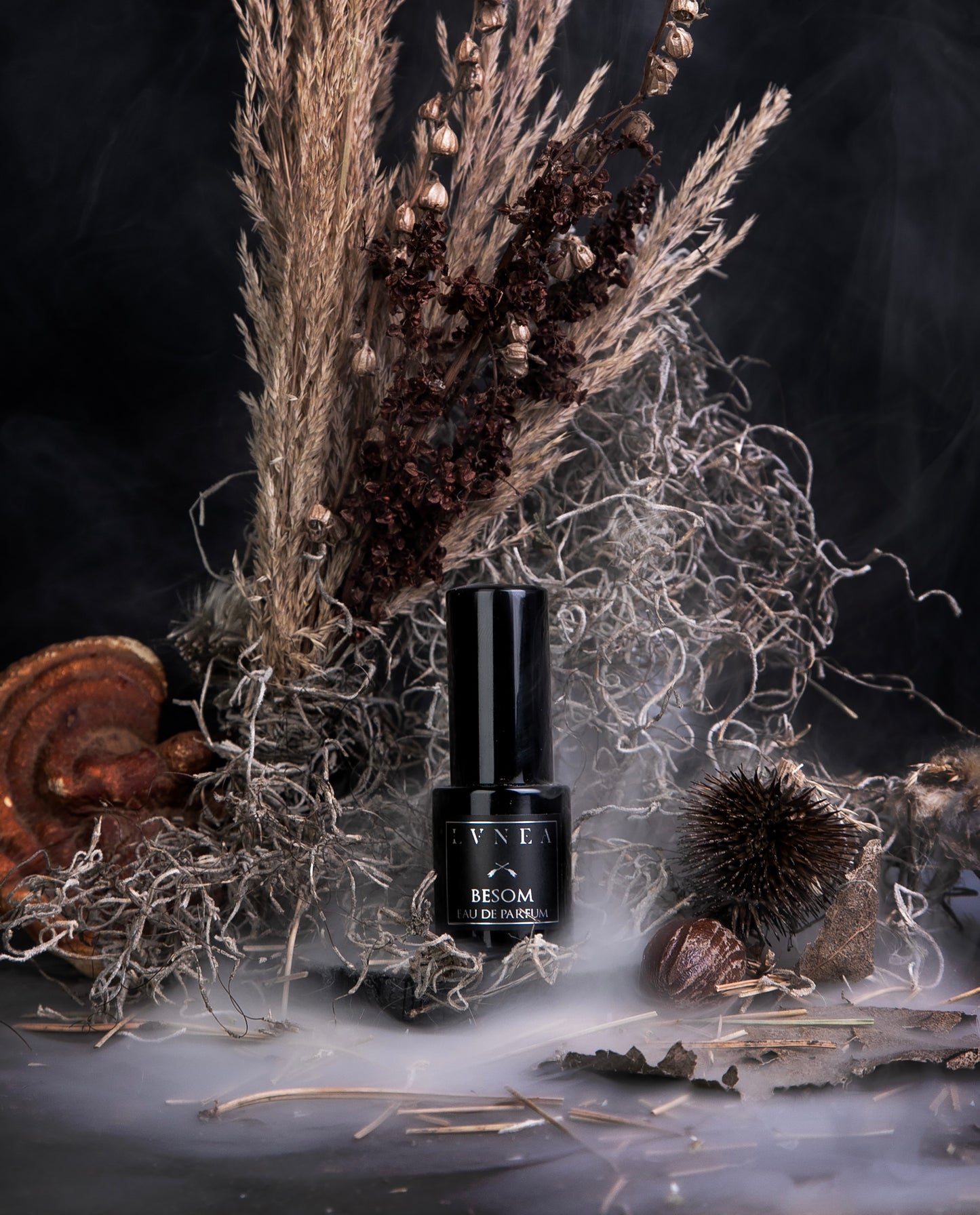 A 15ml black glass bottle of LVNEA's limited-edition eau de parfum "Besom", standing in front of a bouquet of dried grasses and brown botanicals, smoke weaving through the scene.