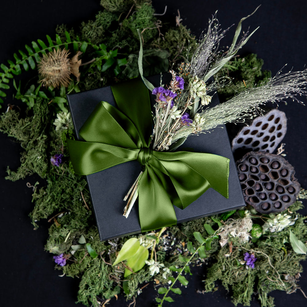 
                      
                        Black cardboard box wrapped in an olive green silk ribbon, sitting atop lush greenery and botanicals. The box contains LVNEA's limited edition THE WITCH'S GARDEN seasonal offerings.
                      
                    