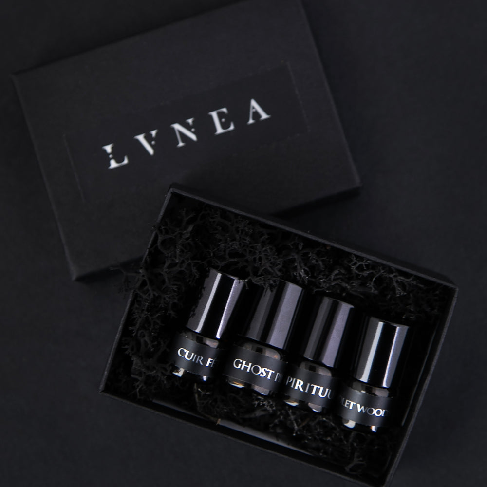 Set of 4 1.25 ml glass sample vials of LVNEA oil perfume. Shown are Cuir Fétiche, Ghost Pine, Spirituum, and Violet Woods. They are in a black box filled with moss, on a black background.