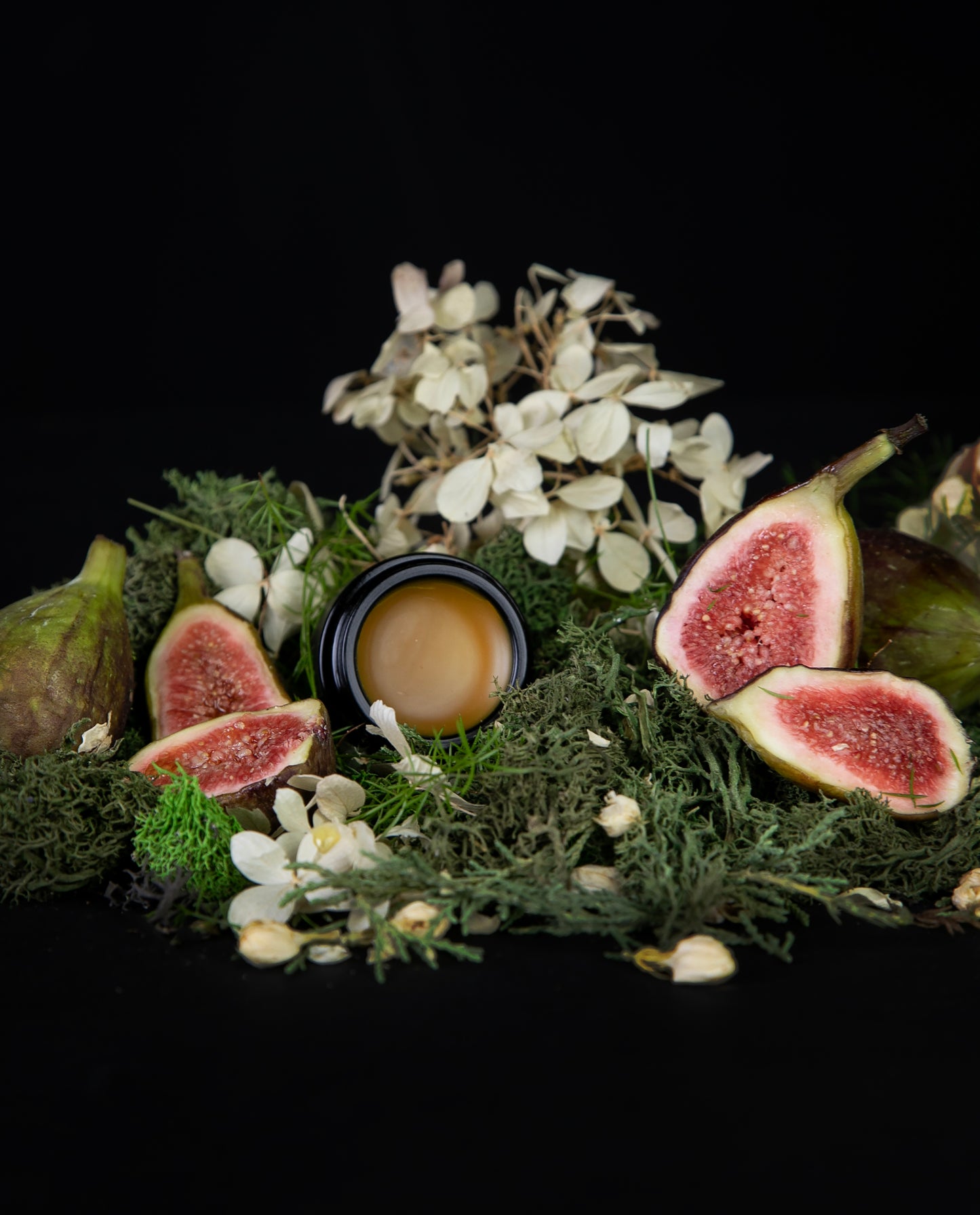 10g black glass pot of LVNEA's 'Jasmine & Fig' solid perfume, lid open to reveal a deep yellow balm. The jar is nestled amongst greenery, white florals, and fresh juicy figs.