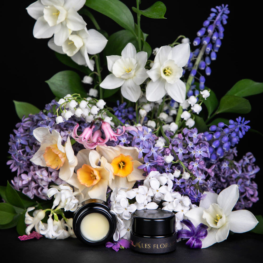 Black glass pot of LVNEA's "Les Floraisons Précieuses" limited edition enfleurage parfum crème, surrounded by lilac, hyacinth, paper whites, lily of the valley, and other spring blooms.