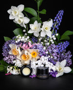 Black glass pot of LVNEA's "Les Floraisons Précieuses" limited edition enfleurage parfum crème, surrounded by lilac, hyacinth, paper whites, lily of the valley, and other spring blooms.