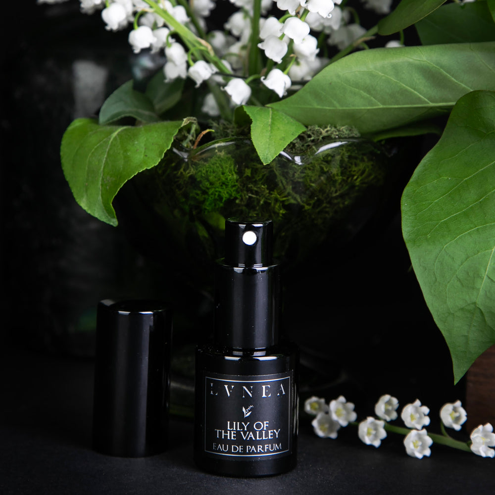 15ml black glass bottle of LVNEA's limited edition Lily of the Valley perfume, against a black background, surrounded by fresh lily of the valley and greenery. The lid of the bottle is open, revealing a spray top