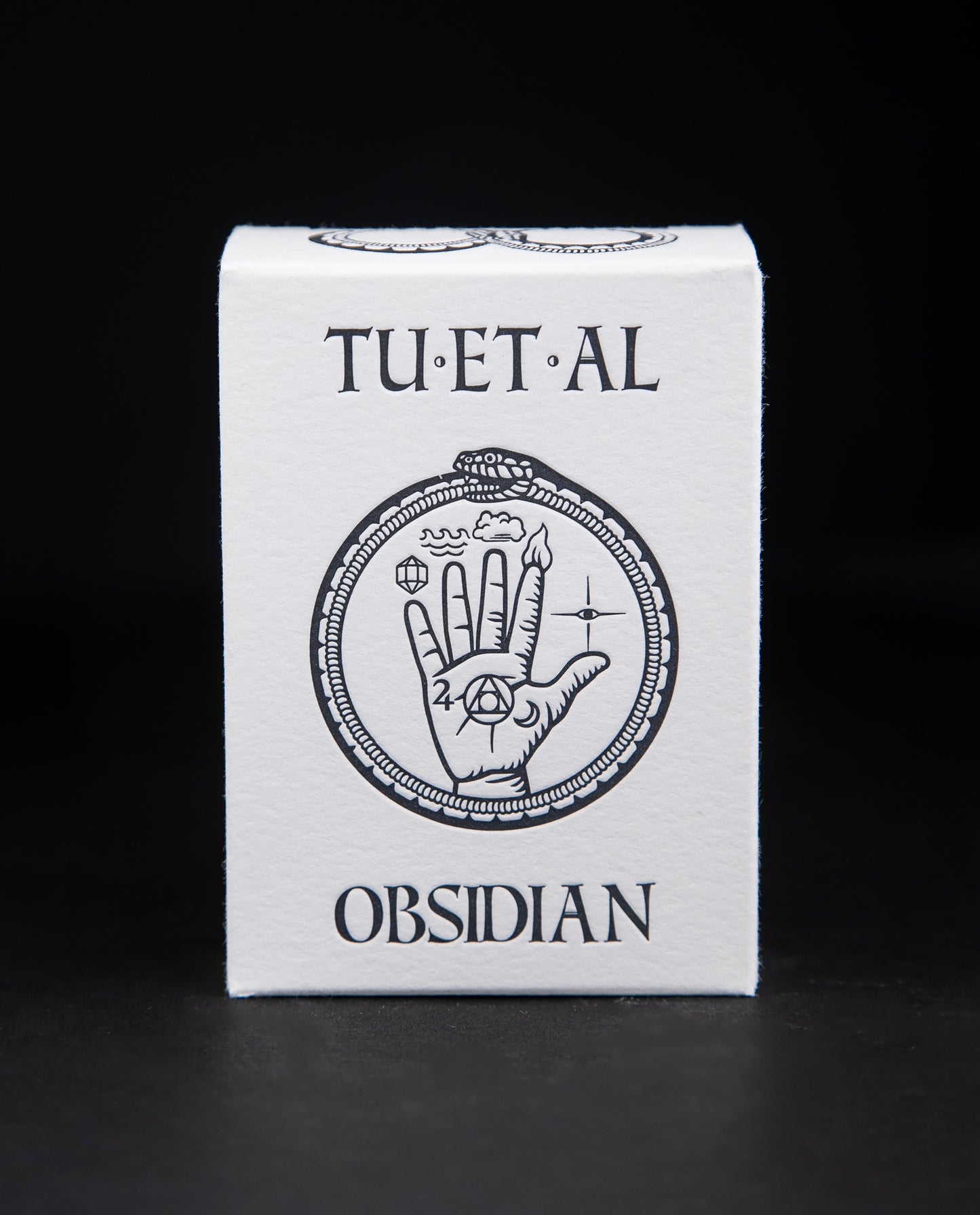 White cardboard soap box containing TU-ET-AL's "Obsidian" soap. There is an illustration of a snake eating it's own tail, and an alchemical hand on front in black.