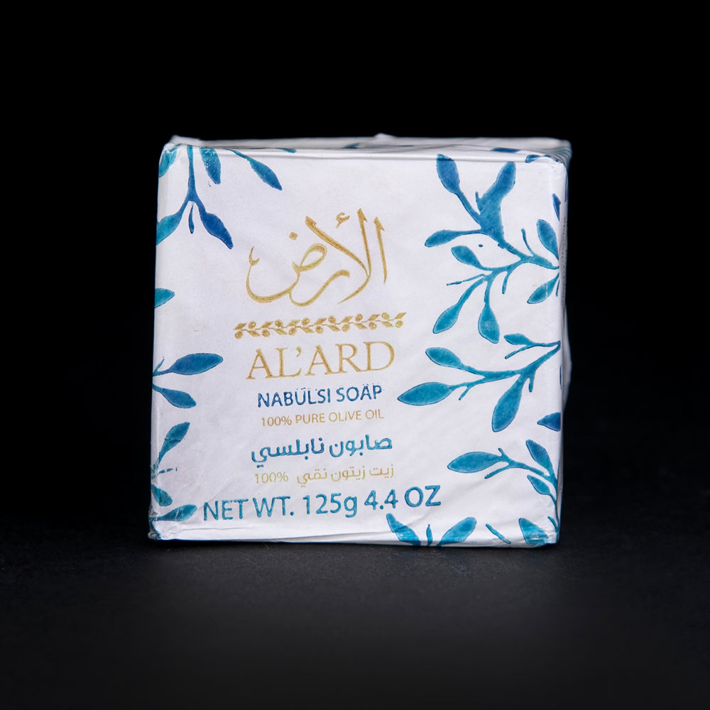 Bar of Palestinian Olive Oil soap wrapped in white wax paper, stamped with blue olive leaves and a gold "Al'Ard" logo