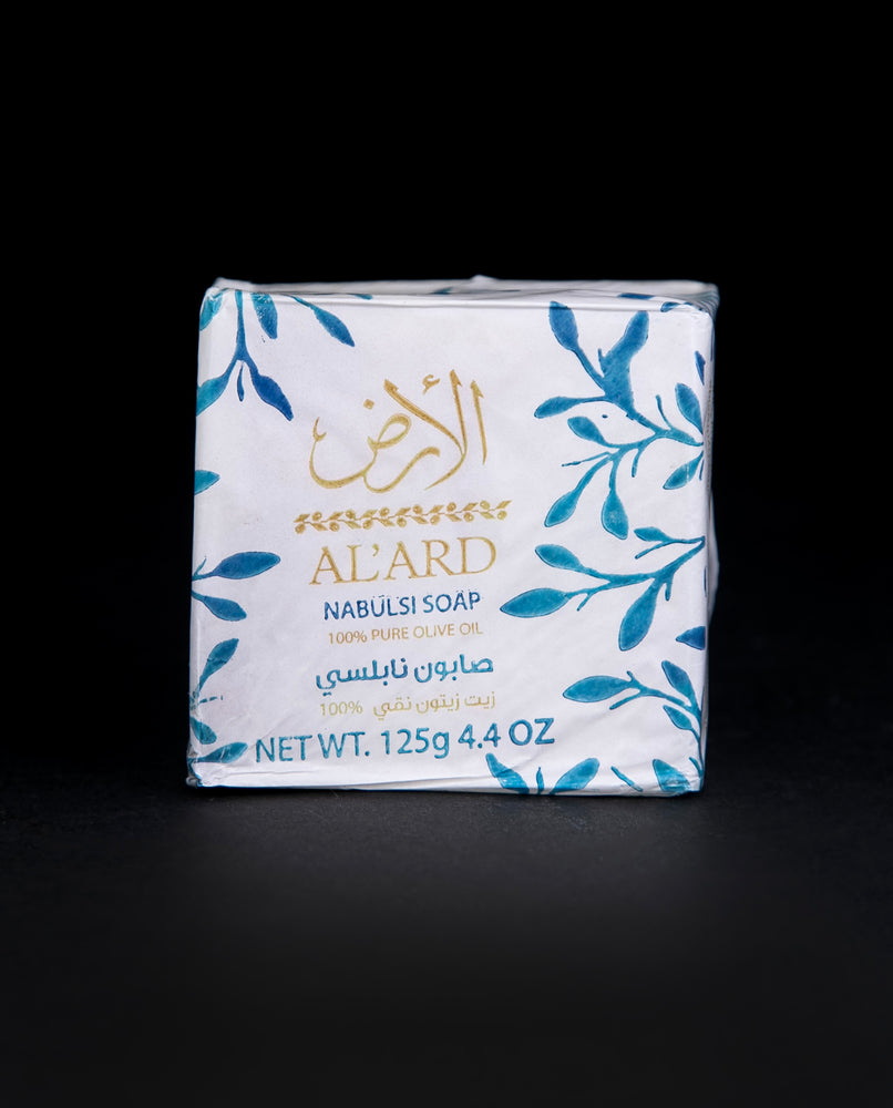 Bar of Palestinian Olive Oil soap wrapped in white wax paper, stamped with blue olive leaves and a gold "Al'Ard" logo