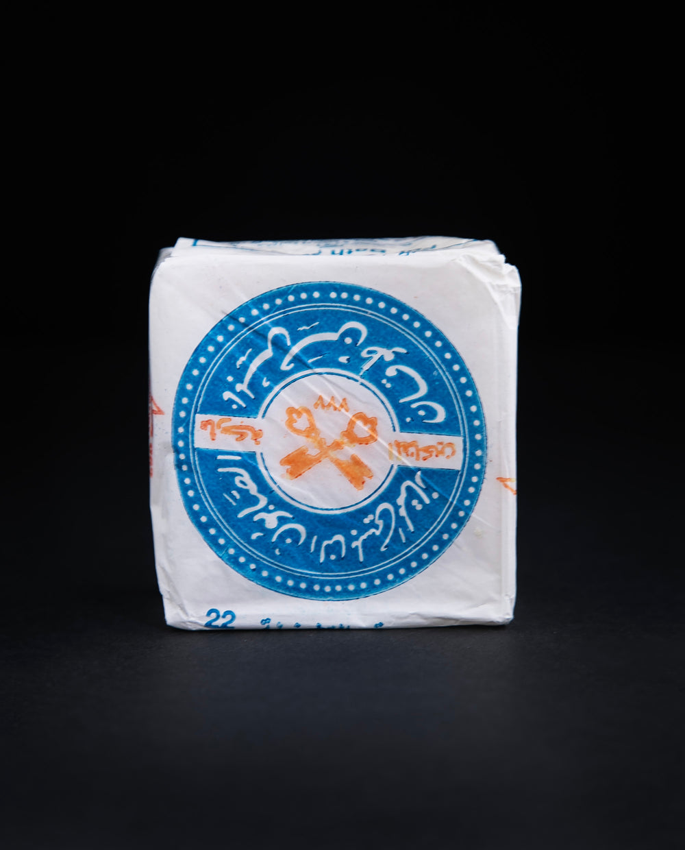 Front view of a bar of Palestinian olive oil soap, wrapped in white wax paper stamped with blue and red 