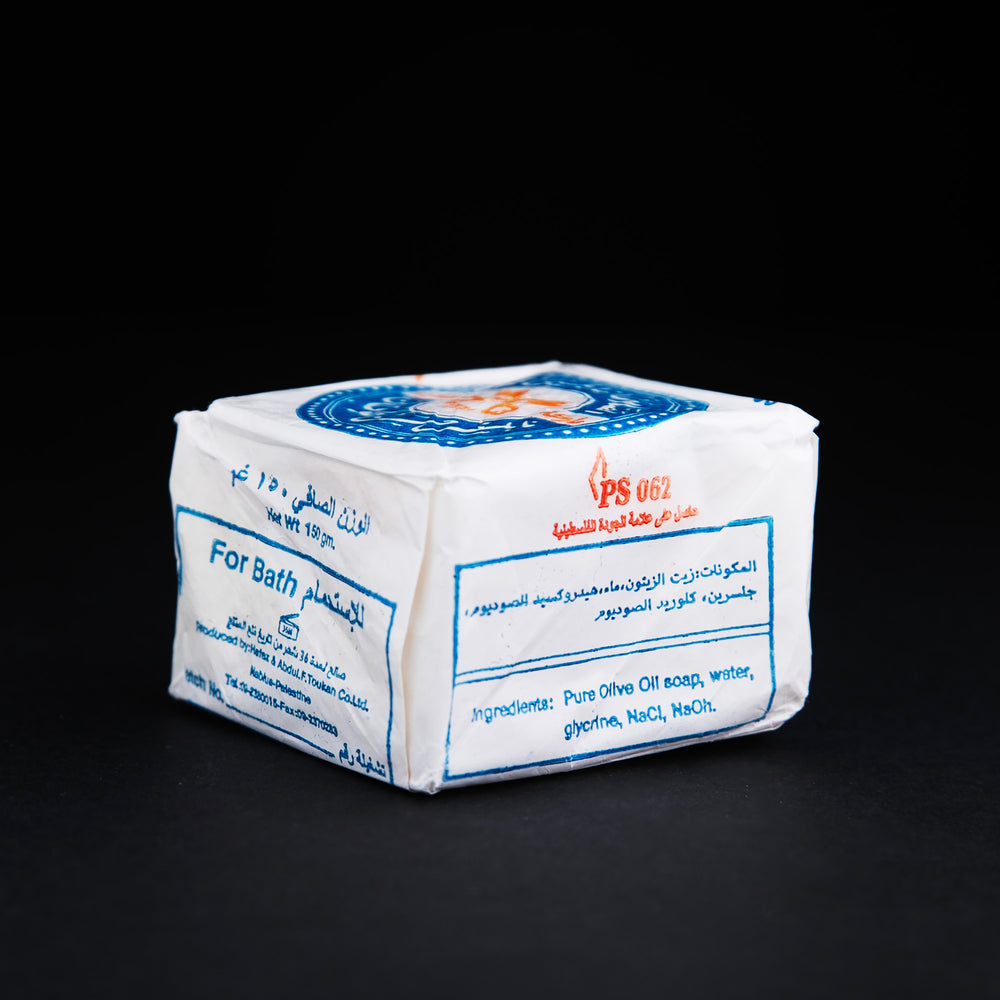 
                      
                        Side view of a bar of Palestinian olive oil soap, wrapped in white wax paper stamped with blue and red "Two Keys" logo
                      
                    