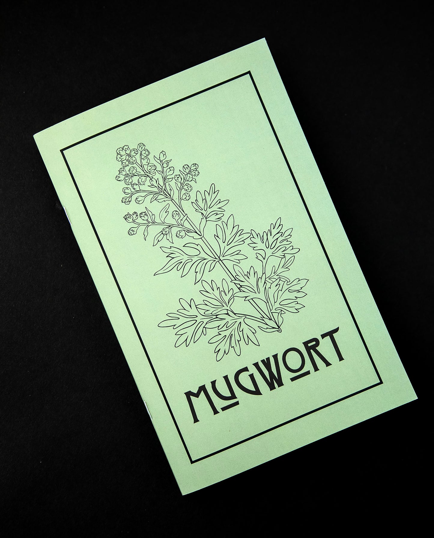 Herbal Revolution's zine about mugwort on black background. The cover is light green, reads "Mugwort" in bold lettering, and features a simple illustration of a mugwort plant.