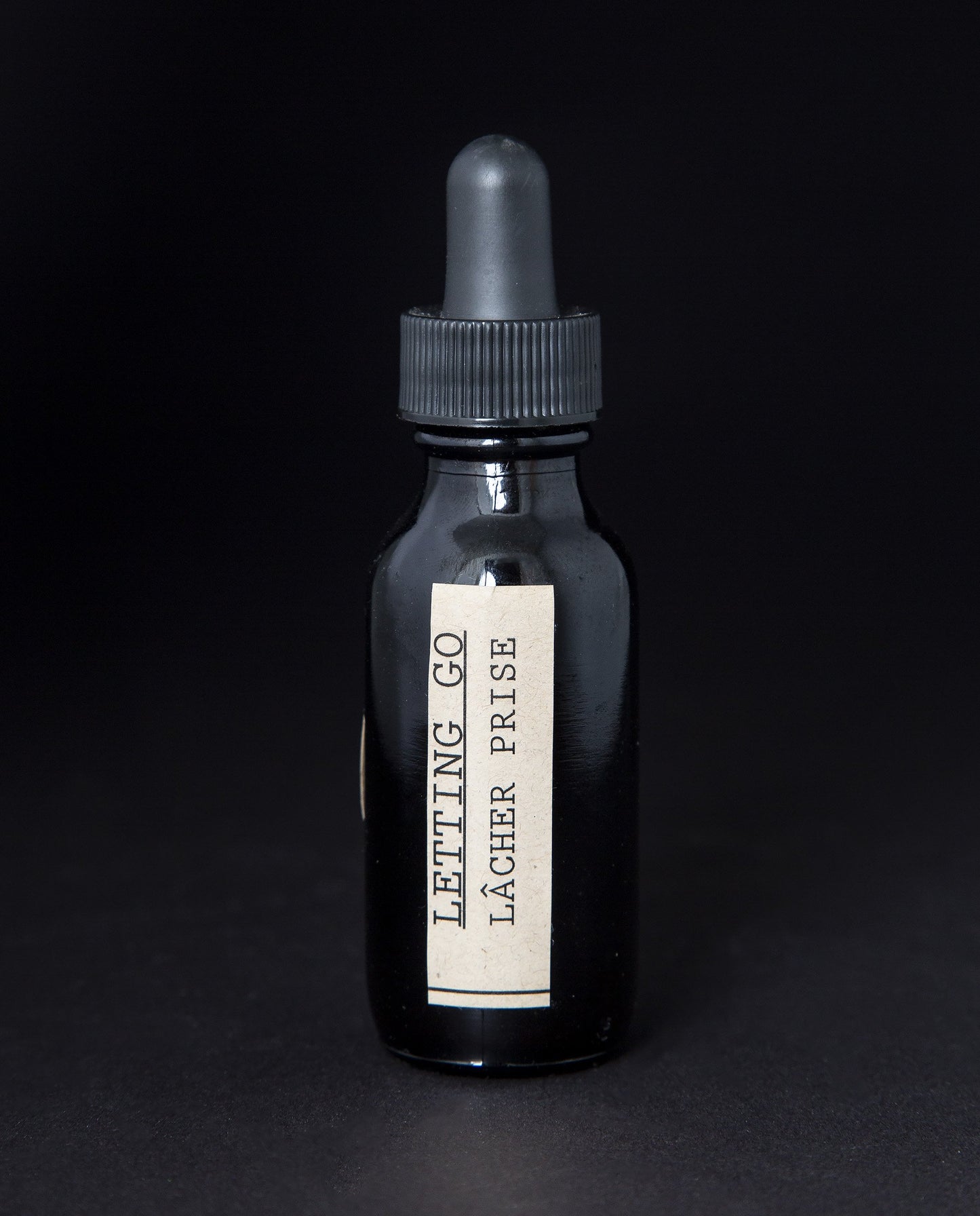 Black glass bottle with dropper top on black background, containing a herbal tincture by blueberryjams. The label is cream coloured and reads "Letting Go / Lâcher Prise".