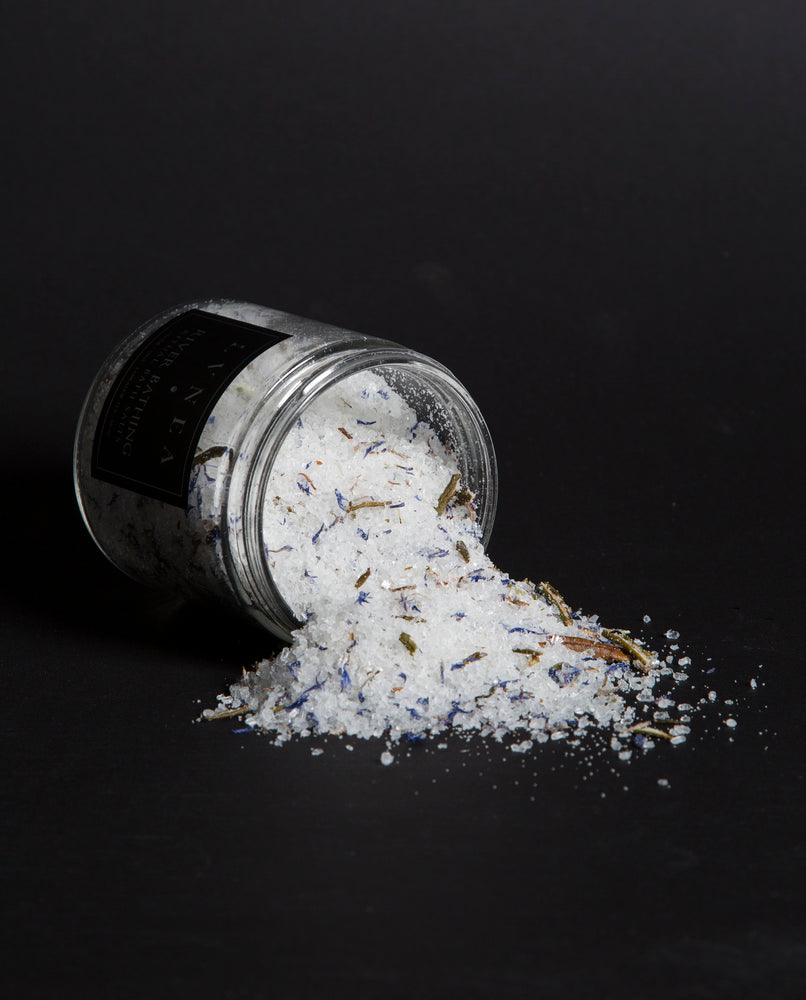 LVNEA'S River Bathing bath salts spilling out of a tipped over 16 ounce clear glass jar. There are blue cornflower petals, labrador tea buds, and usnea in visible in the salt mix.