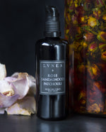 Black bottle of LVNEA's Rose, Sandalwood and Patchouli body serum standing next to large clear glass jug filled with amber-coloured oil